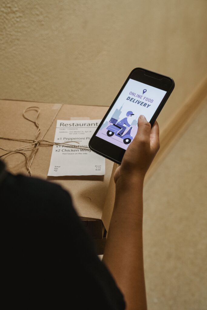 A person holding a phone displaying an online food delivery app next to a pizza box indoors.